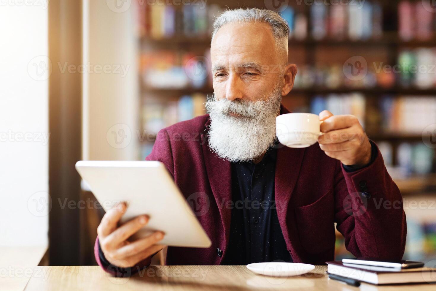 Wealthy grey-haired elderly man in burgundy jacket drinking coffee and using digital tablet, checking his email or reading book while resting at cafe, empty space. Retirement lifestyle concept photo