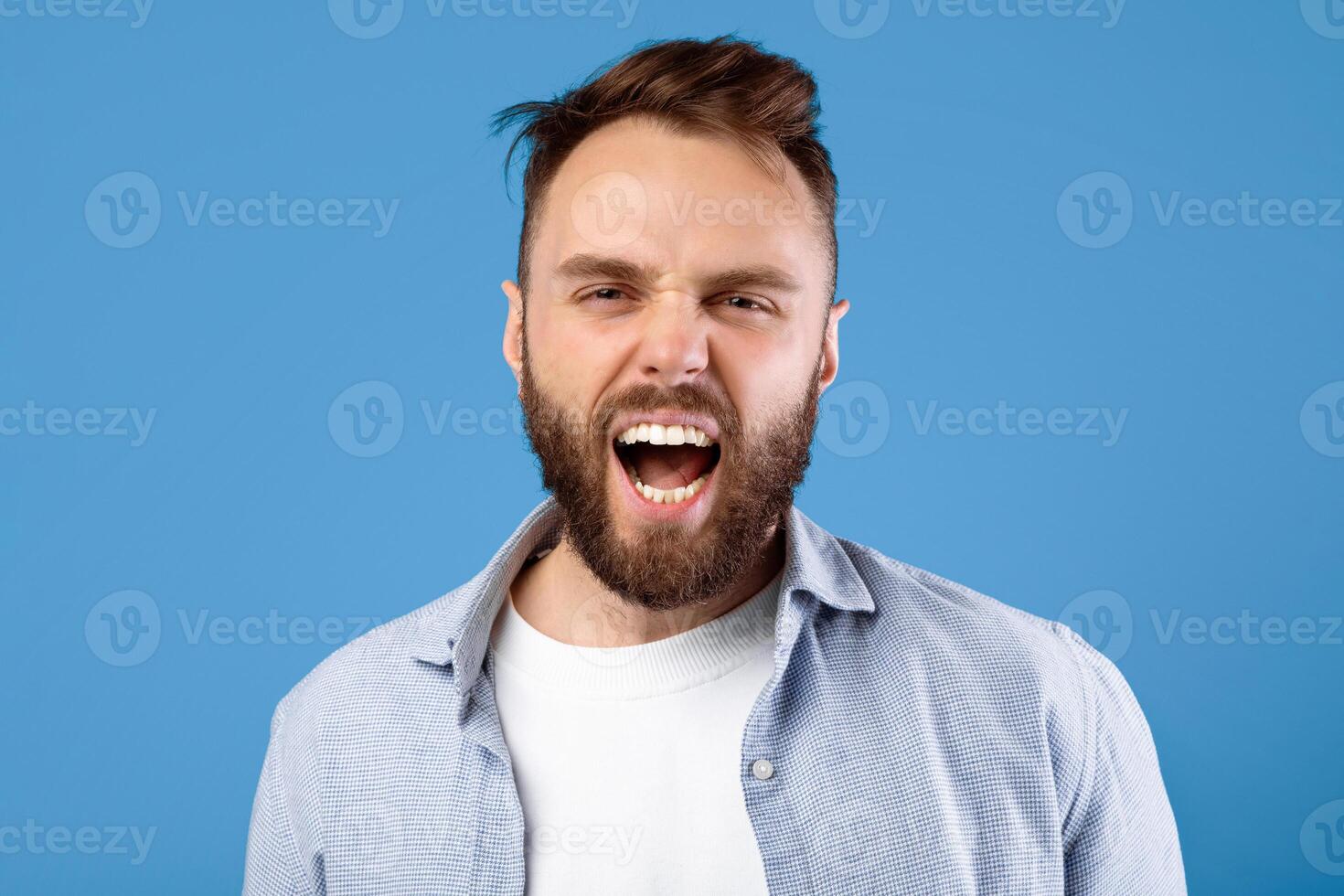 Enraged bearded guy shouting in anger, expressing his aggression over blue studio background. Emotional millennial man yelling, feeling furious or annoyed. Negative human emotions concept photo