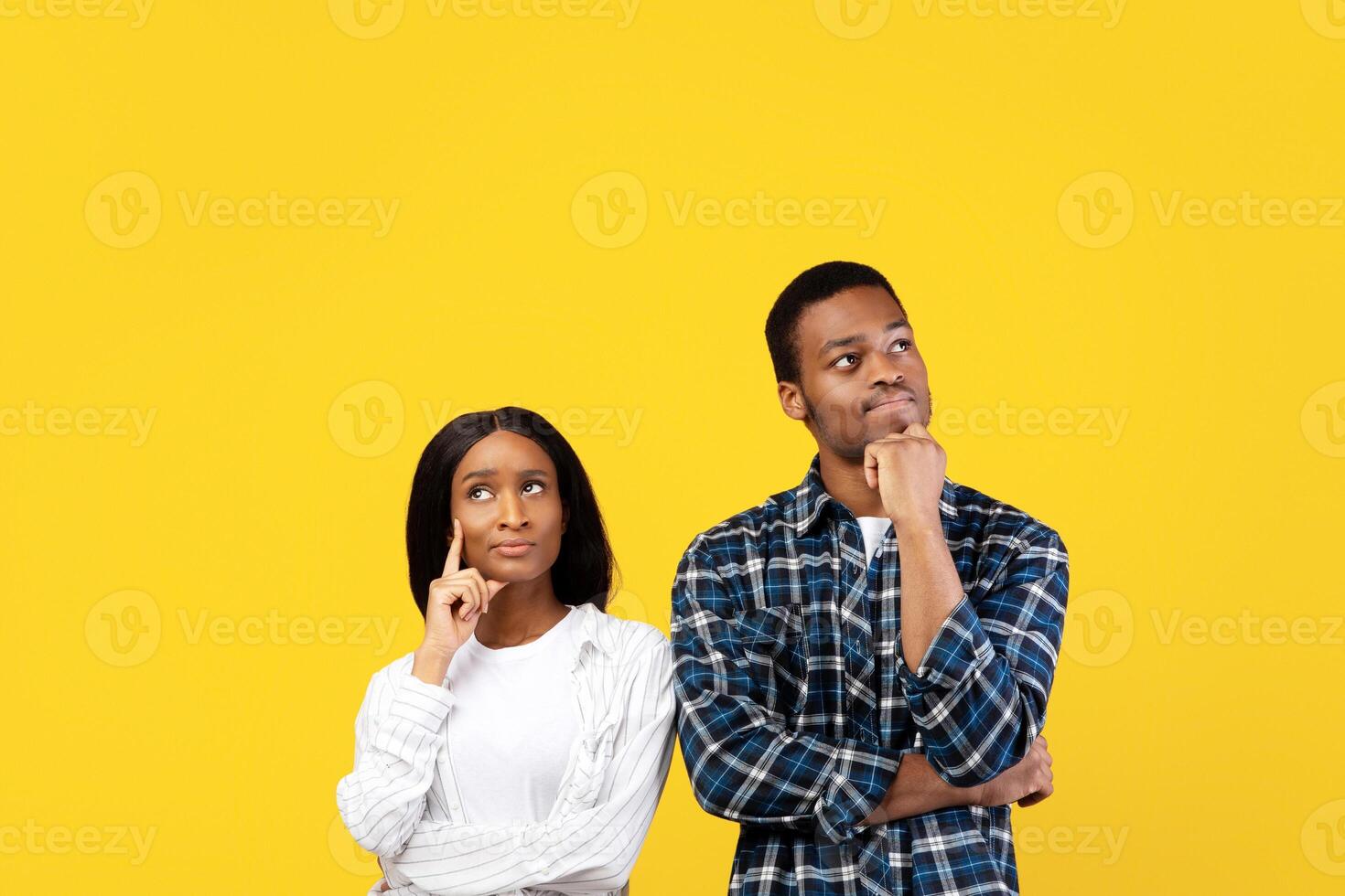 Serious confident couple of two people interested, thinking. Pensive young african american husband and wife, stand under empty space and touch chin, isolated on yellow background, studio shot photo