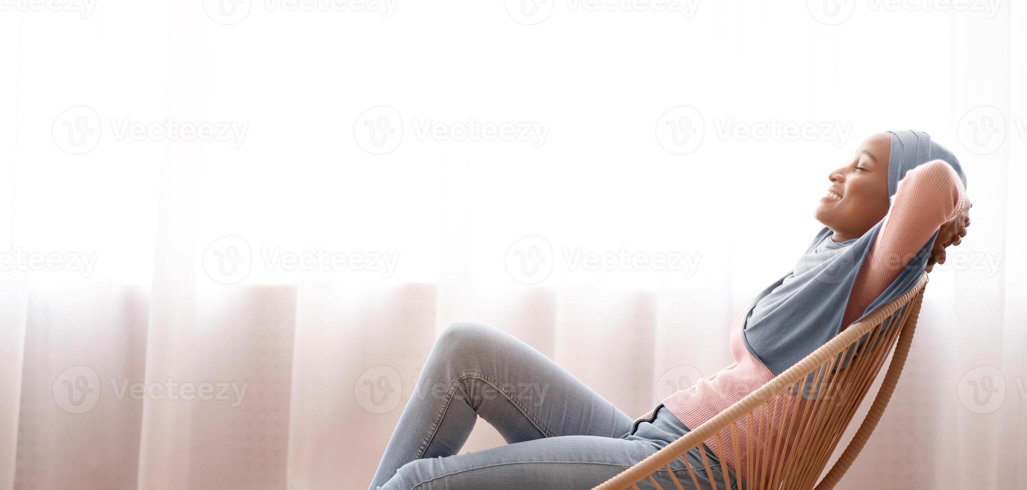 Side view of happy Muslim woman in hijab relaxing in comfortable armchair, resting her feet on knitted pouf, copy space. Young lady in Arabic headscarf spending lazy peaceful weekend at home photo