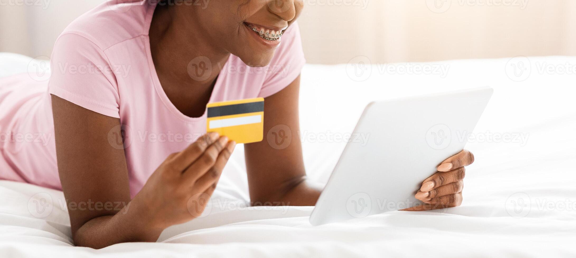 Happy african american lady shopping from home, using digital tablet while laying on bed, copy space. Joyful black woman holding credit card, using mobile application for e-commerce on her pad photo