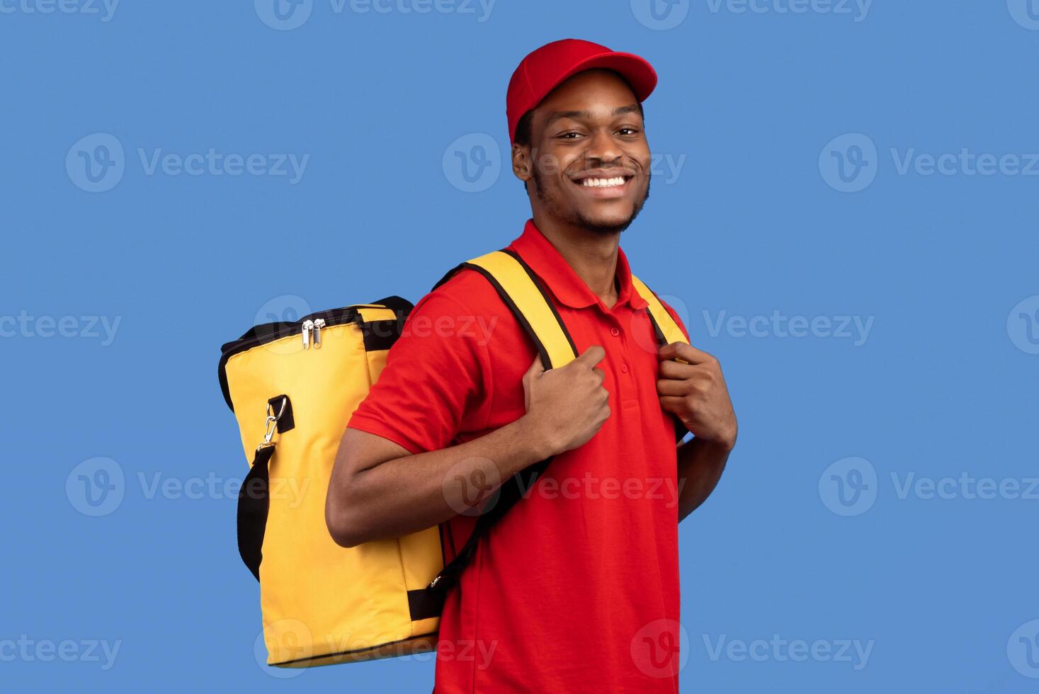 Portrait of cheerful smiling african american deliveryman wearing red uniform, cap and yellow thermal refrigerator backpack looking at camera isolated on blue background, posing at studio photo