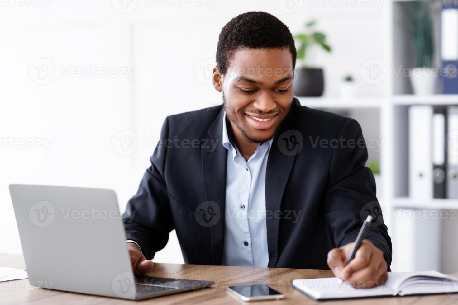 Smiling african american entrepreneur having online training in office, using laptop, taking notes, copy space. Cheerful black businessman working with computer and writing in notepad photo