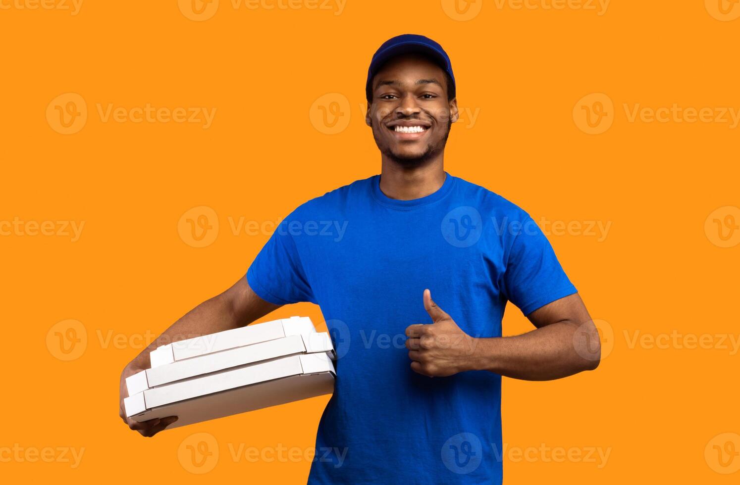 Best Fast Food Delivery. Portrait of smiling african courier wearing blue cap and uniform holding pizza boxes from restaurant and showing thumb up gesture sign, isolated on orange studio background photo