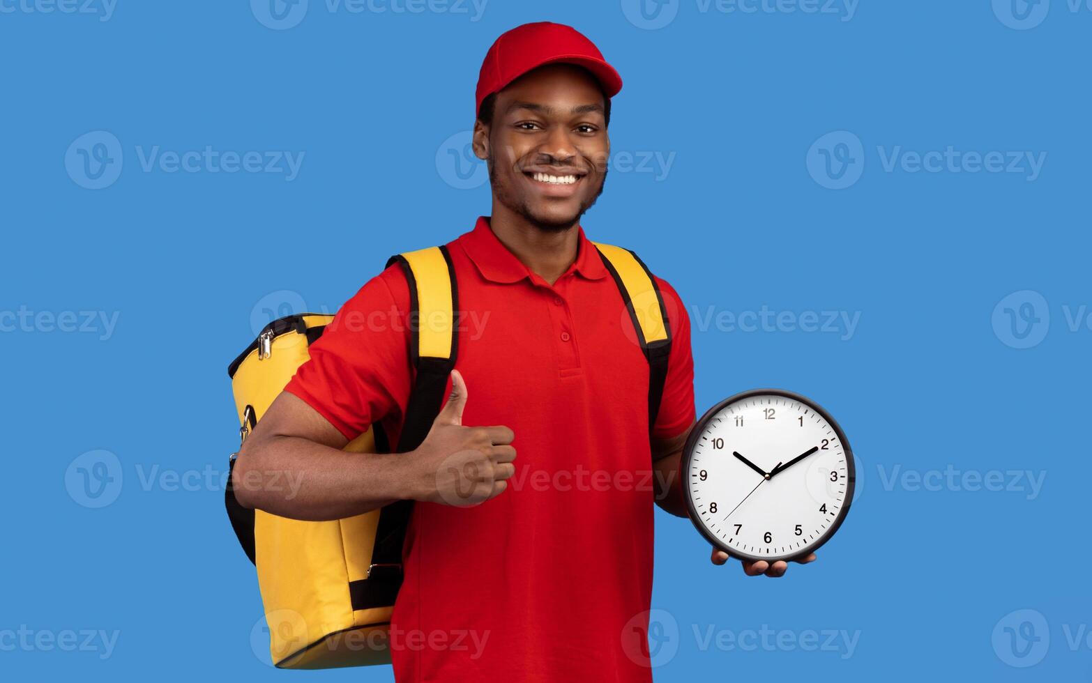 Fast Delivery Service Concept. Portrait of confident smiling black male courier wearing red uniform and thermo backpack bag holding clock and showing thumbs up gesture isolated on blue background photo
