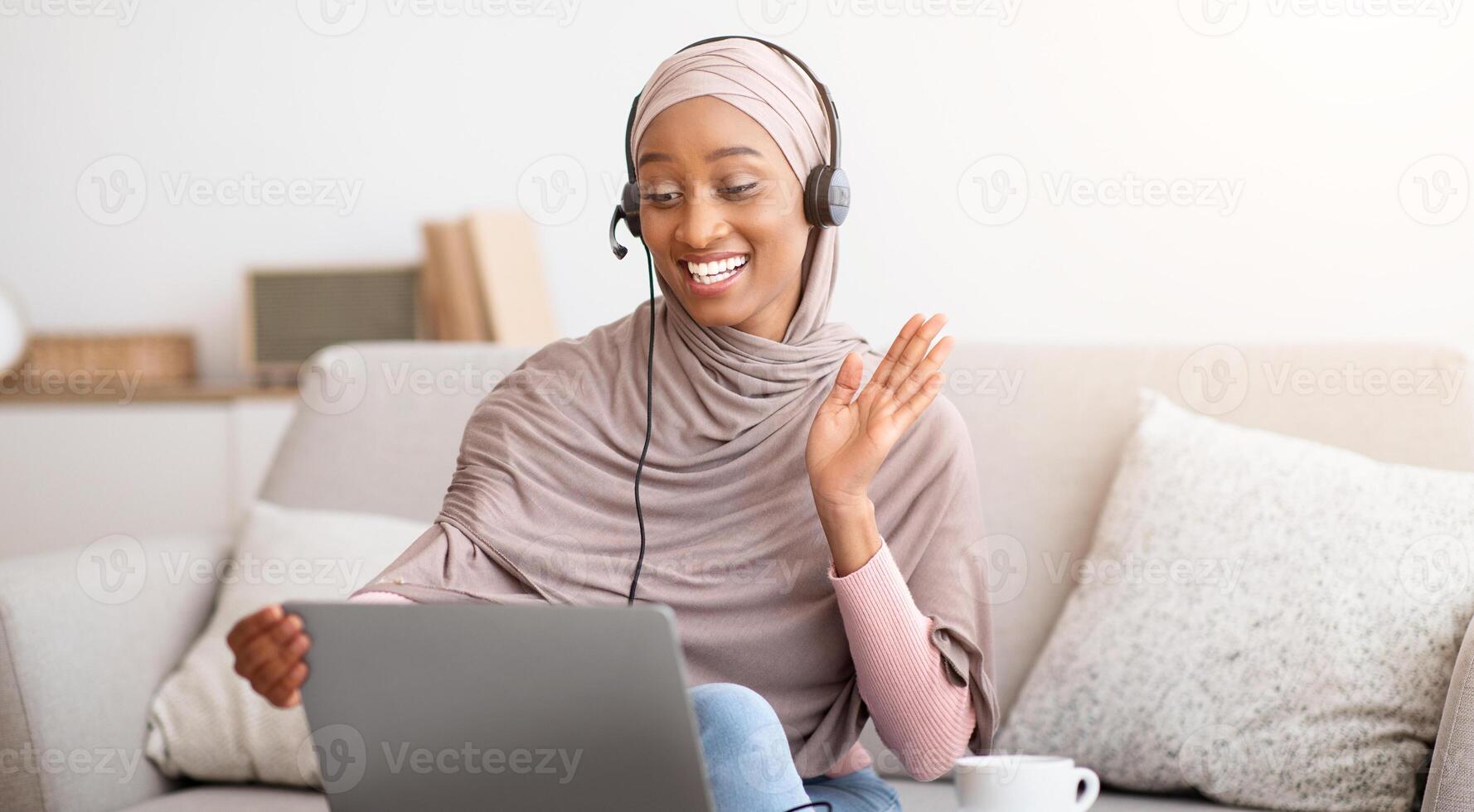Happy black Muslim lady in hijab and headset making call on laptop at home, waving at screen. Lovely young Arab woman enjoying online communication, participating in web conference photo