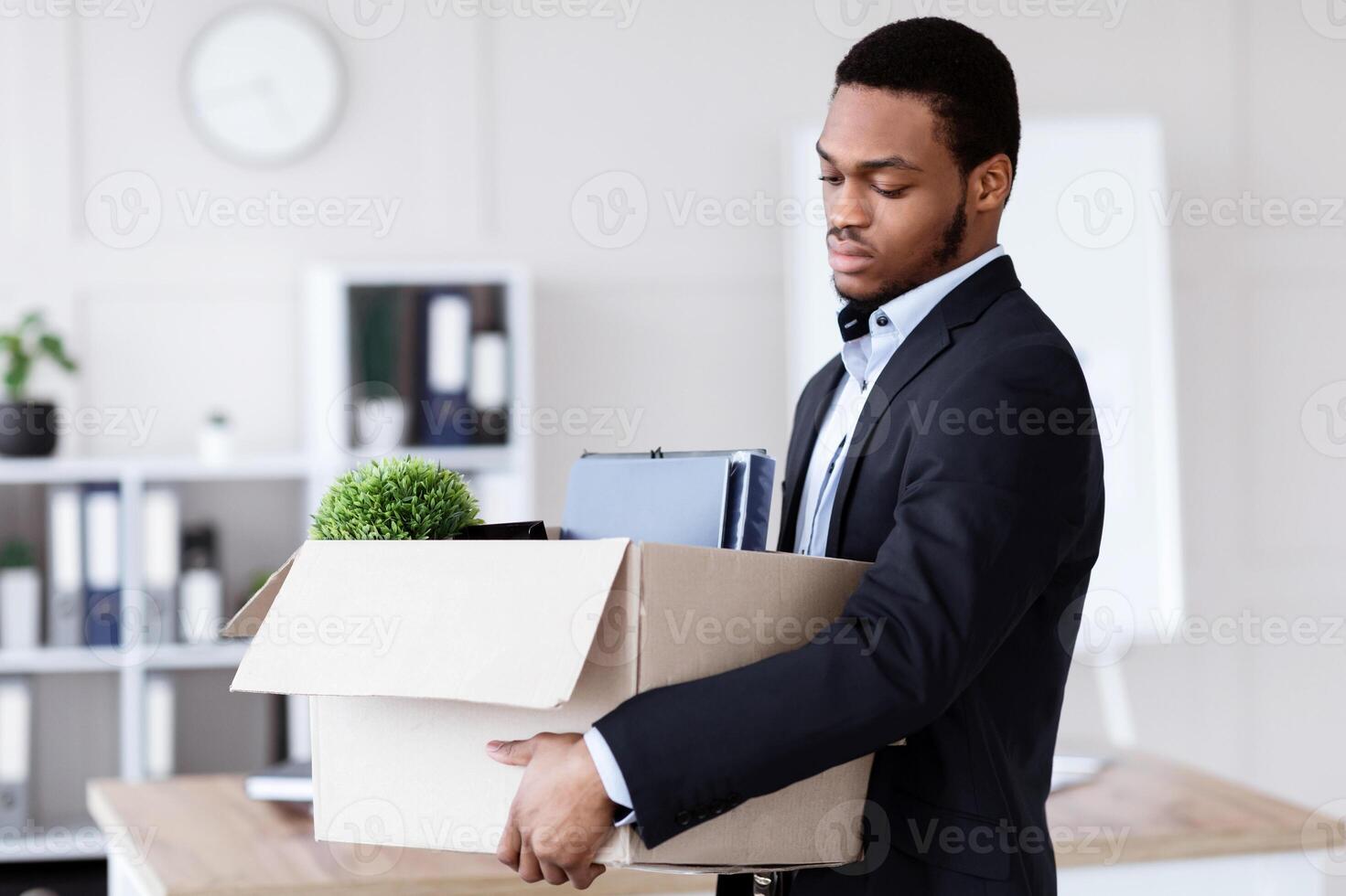 Upset black man got fired, standing in office and holding box with his belongings, copy space. Terminated african american guy in suit lost his job, packing his working stuff before leaving workplace photo