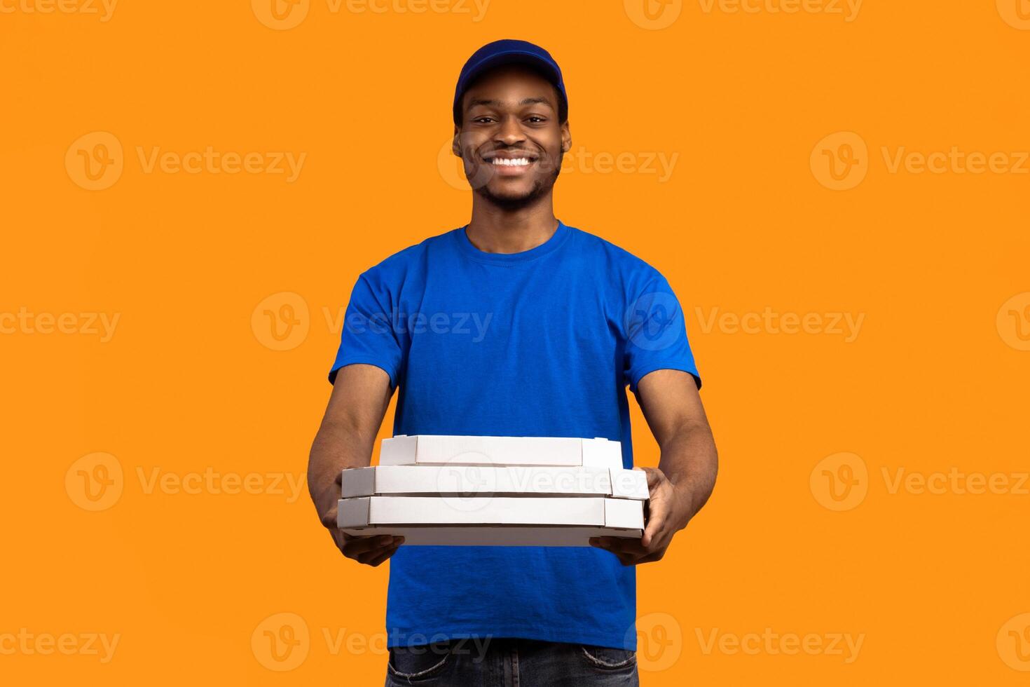 Fast Food Delivery. Portrait of smiling african courier wearing blue cap and uniform holding pizza boxes from restaurant and giving it to camera, standing isolated on orange yellow studio background photo