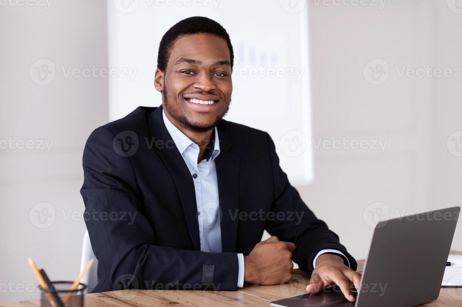 Businessman At Work. Happy african american young businessman in black suit sitting in front of laptop and looking at camera, sitting at desk in his modern office, working, copy space photo
