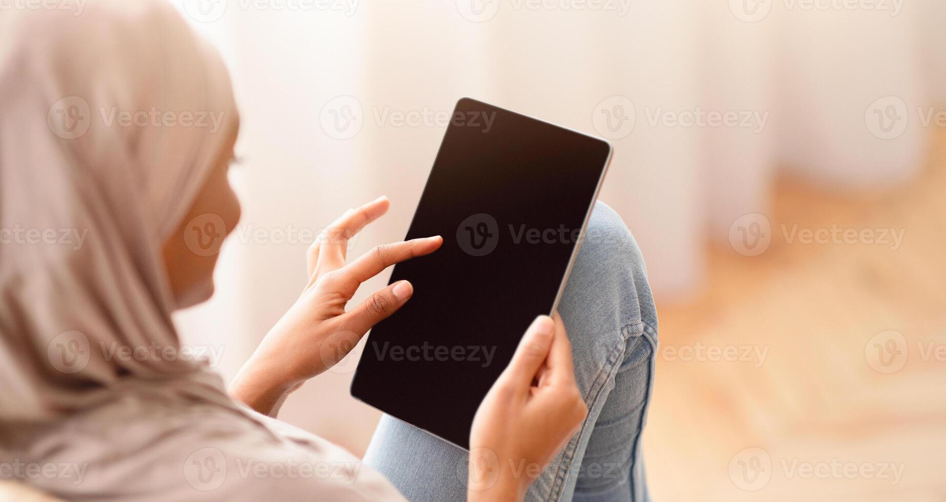 Black Arab woman in hijab using tablet computer with empty screen while sitting in cozy armchair at home, mockup for website design. Muslim lady with touch pad working or studying online photo