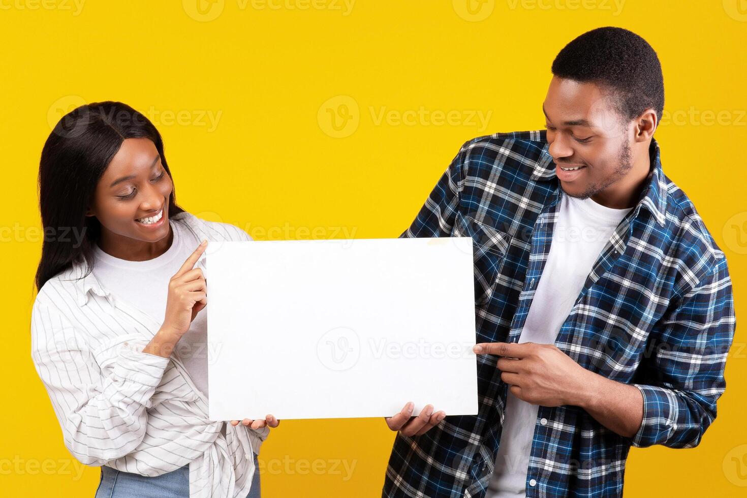 Wow, banner ad, interesting proposal and cool offer. Happy young african american students male and lady in casual holds banner with blank space and look at it, isolated on yellow background photo
