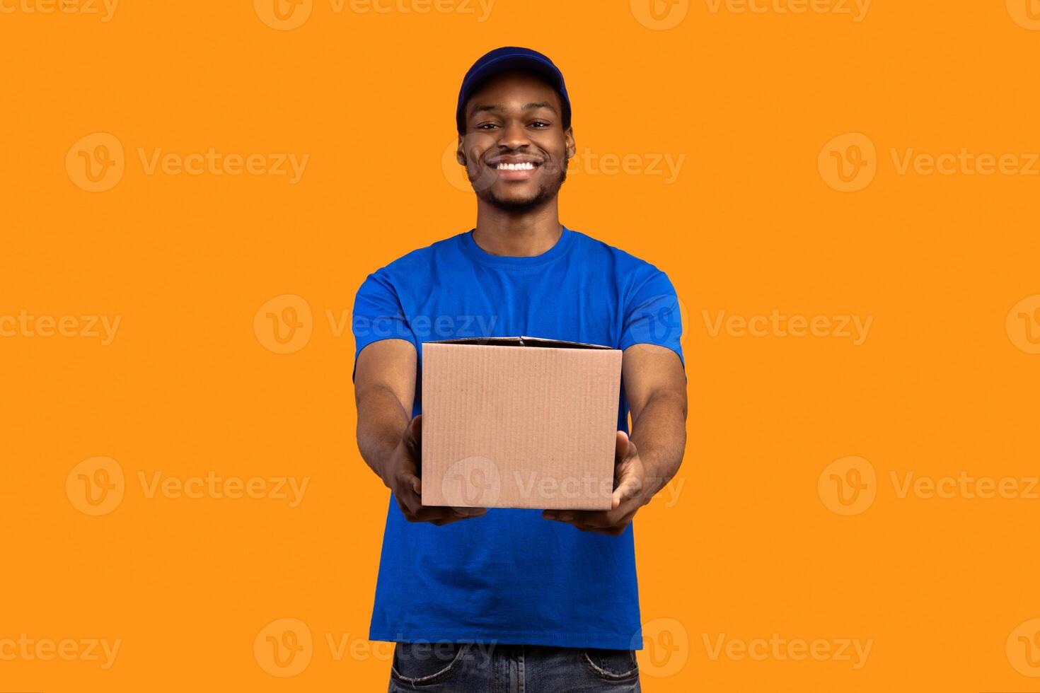 Best Service Concept. Smiling black delivery man wearing blue cap and unform holding cardboard box, showing and giving it to camera standing isolated over yellow orange studio background photo