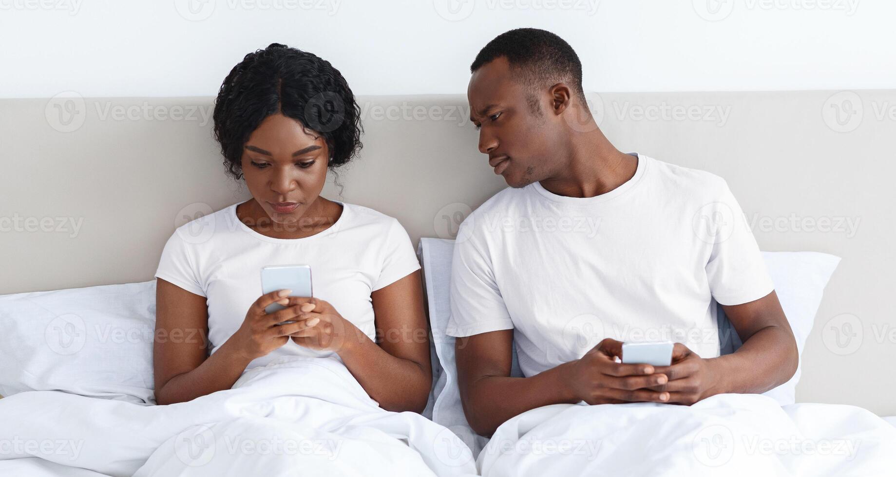 Black guy eavesdropping on his girlfriend using mobile phone, looking at her smartphone screen. African american young couple sitting in bed and using mobile phones. Distrust in relationships photo