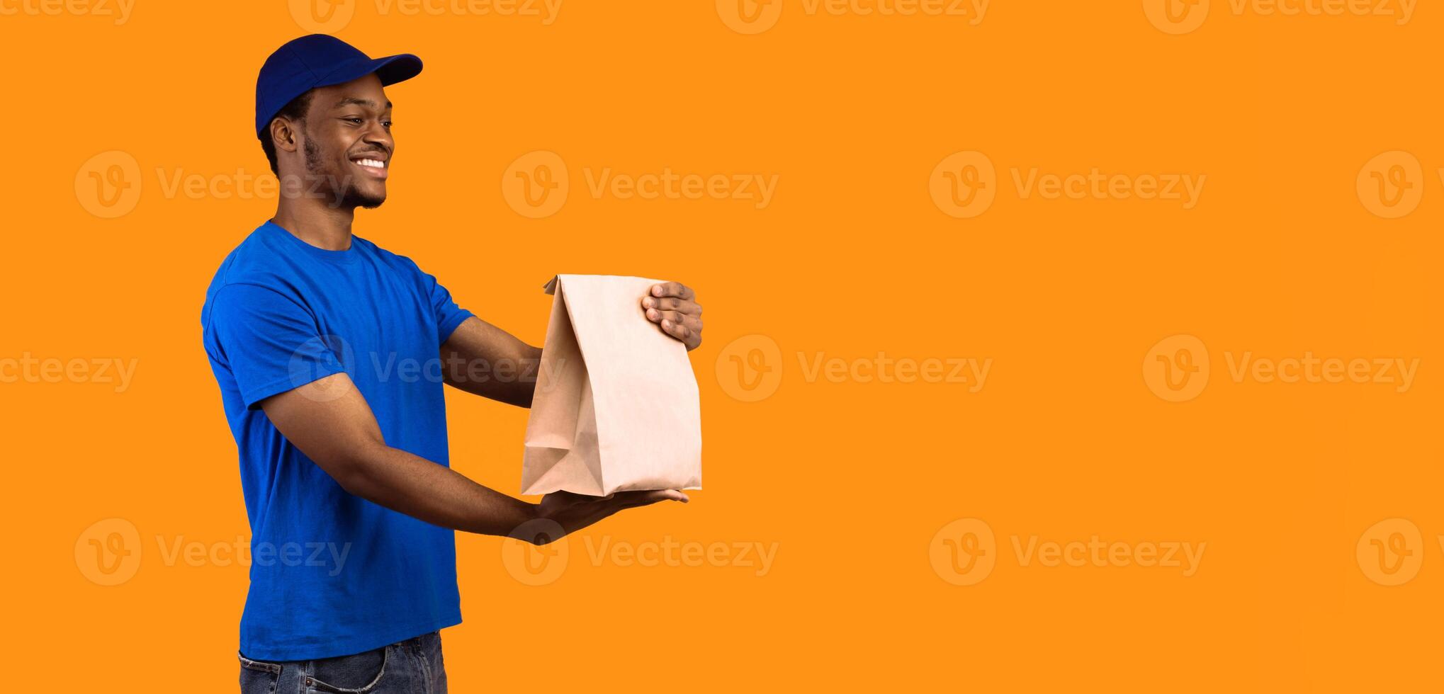Fast Delivery. Side view of smiling african american delivery guy in blue cap holding and giving craft paper bag from restaurant with products to the side with copy space, orange background photo