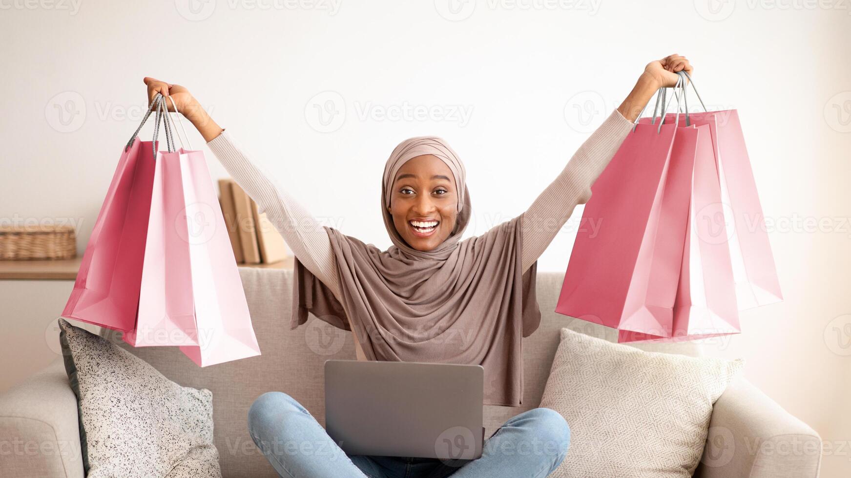 Cheerful black lady in hijab shopping on internet from laptop, lifting paper bags up, feeling excited at home. Happy Muslim woman in headscarf buying goods in web store, ordering online photo