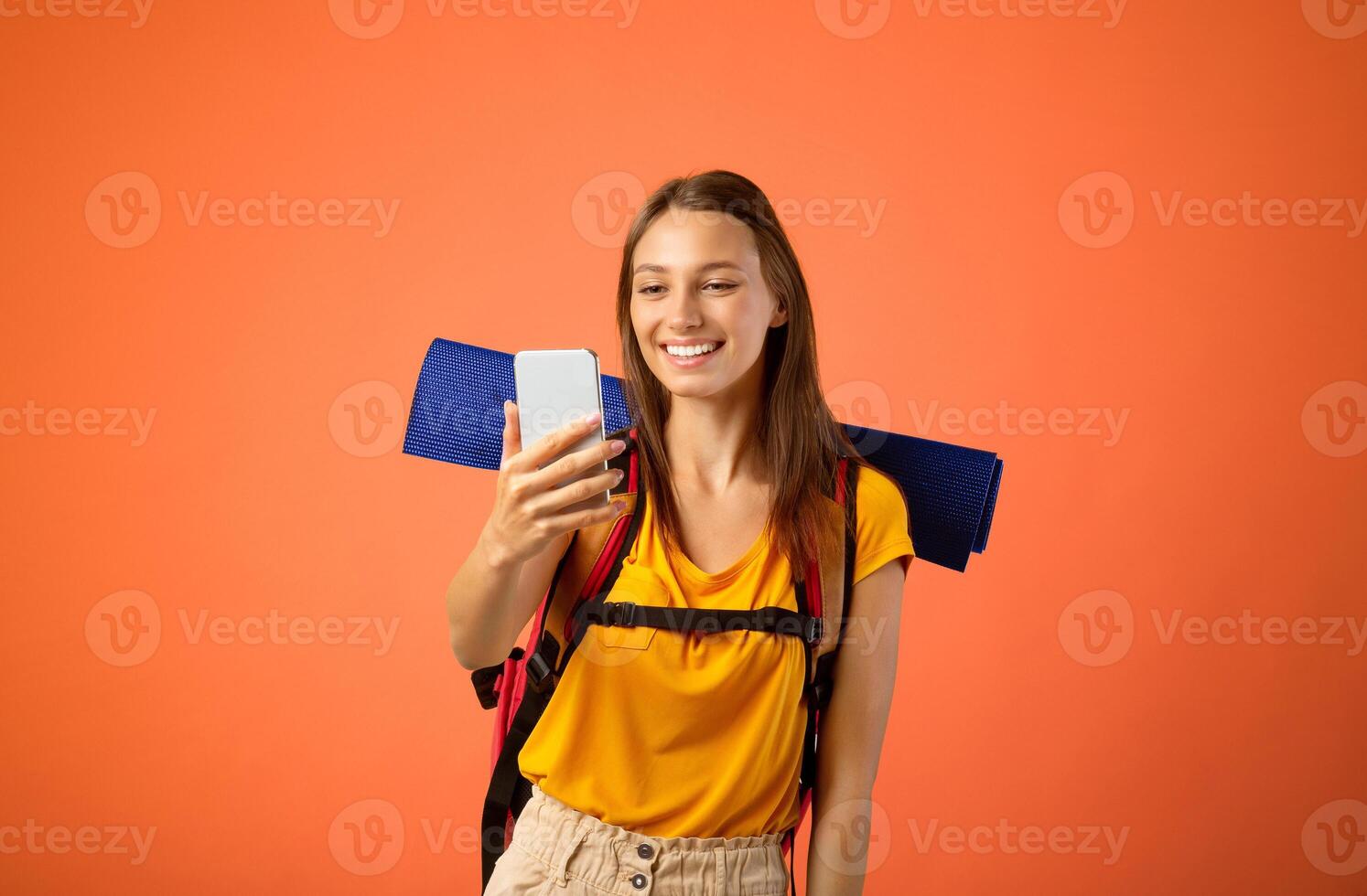Happy young woman with backpack and mat holding mobile phone, having call, copy space, yellow studio background. Female traveller using smartphone, chatting with friends while travelling photo
