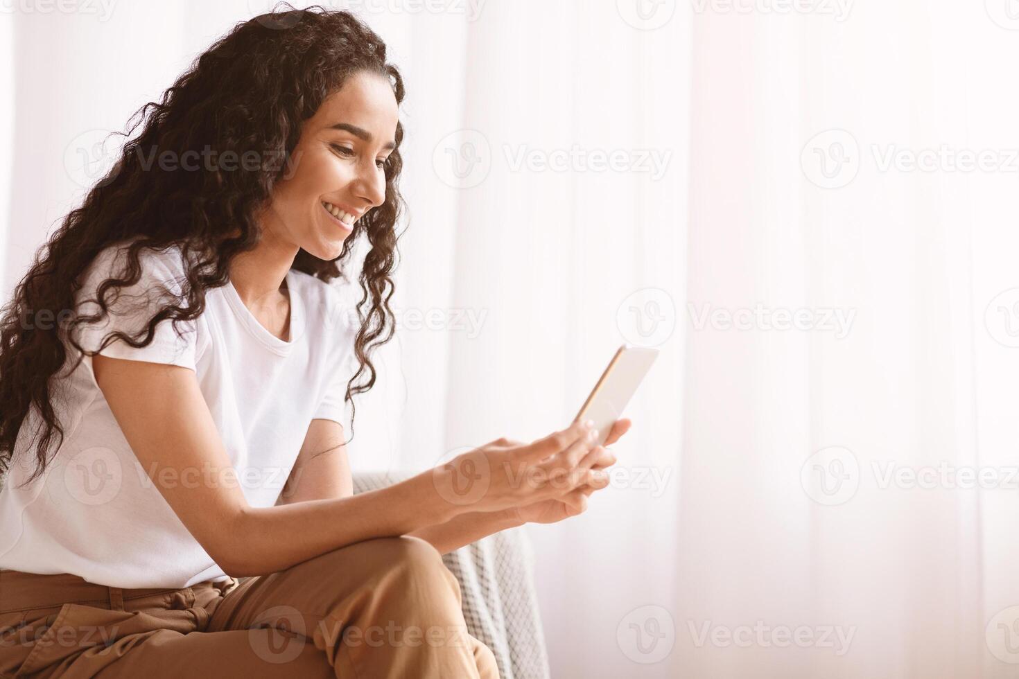 Beautiful smiling young woman with dark curly hair sitting by window and using mobile phone, checking new exciting application for dating, home interior, side view, copy space photo
