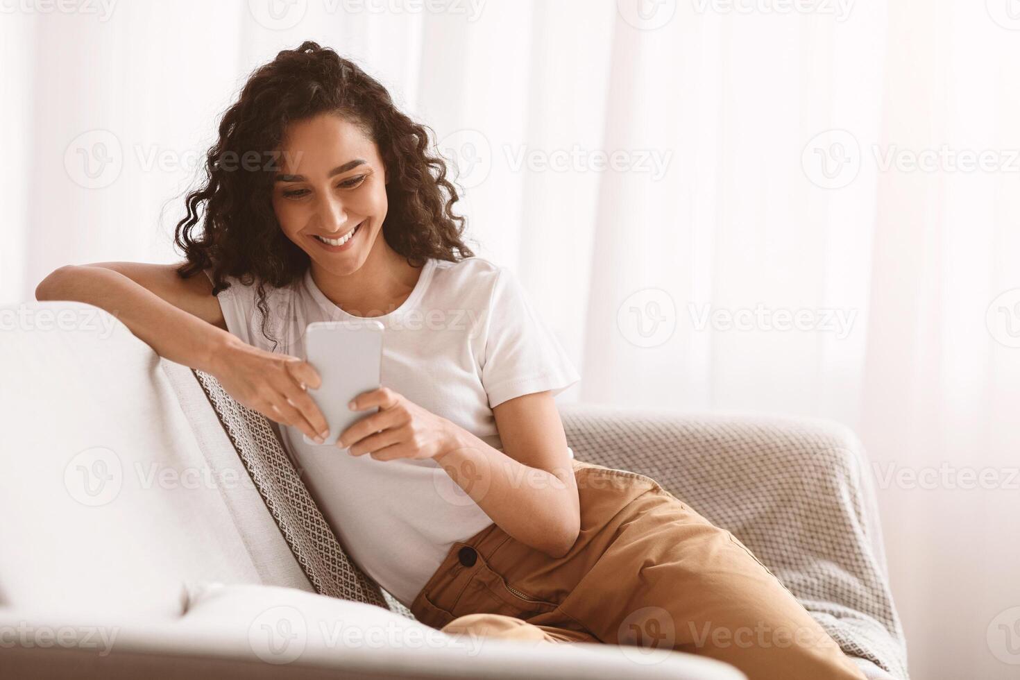Happy beautiful young woman with dark curly hair sitting by window in living room and using mobile phone, checking new exciting application for dating, home interior, side view, copy space photo