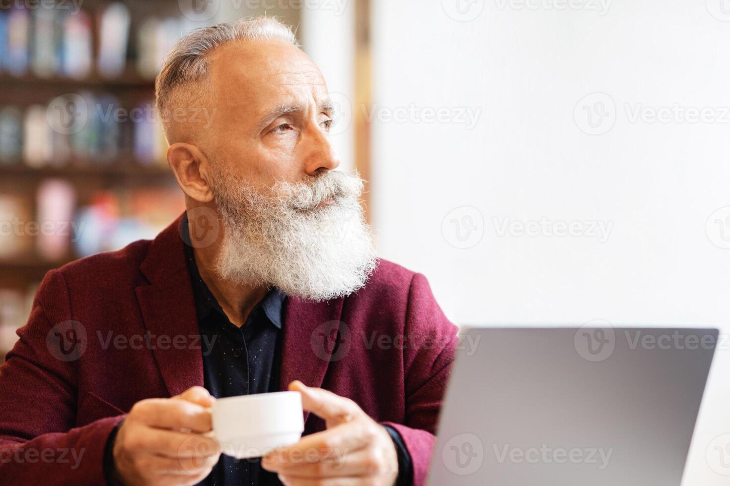 Rich senior man in stylish suit drinking coffee at cafe, sitting in front of modern laptop and looking at copy space. Elderly man using wi-fi while enjoying herbal tea, websurfing or looking for job photo