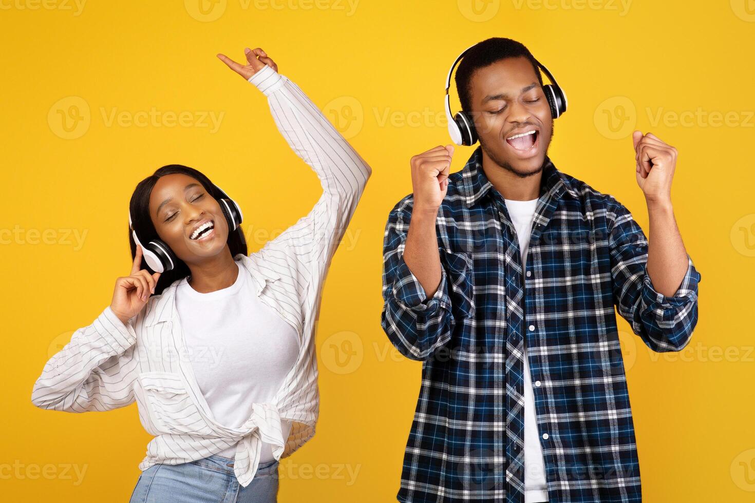 Free time and fun together. Happy young african american man and woman in casual and headphones dancing and singing to favorite music, isolated on yellow background, studio shot, free space photo