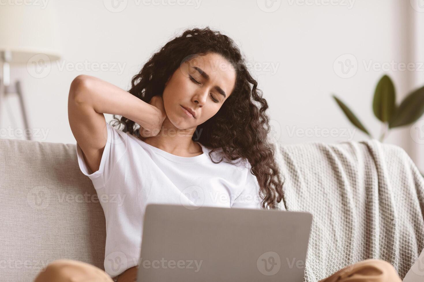 Brunette woman having pain in neck while working with laptop at home, copy space. Dark-haired curly lady rubbing her neck, sitting on couch with laptop on her knees, feeling uncomfortable photo