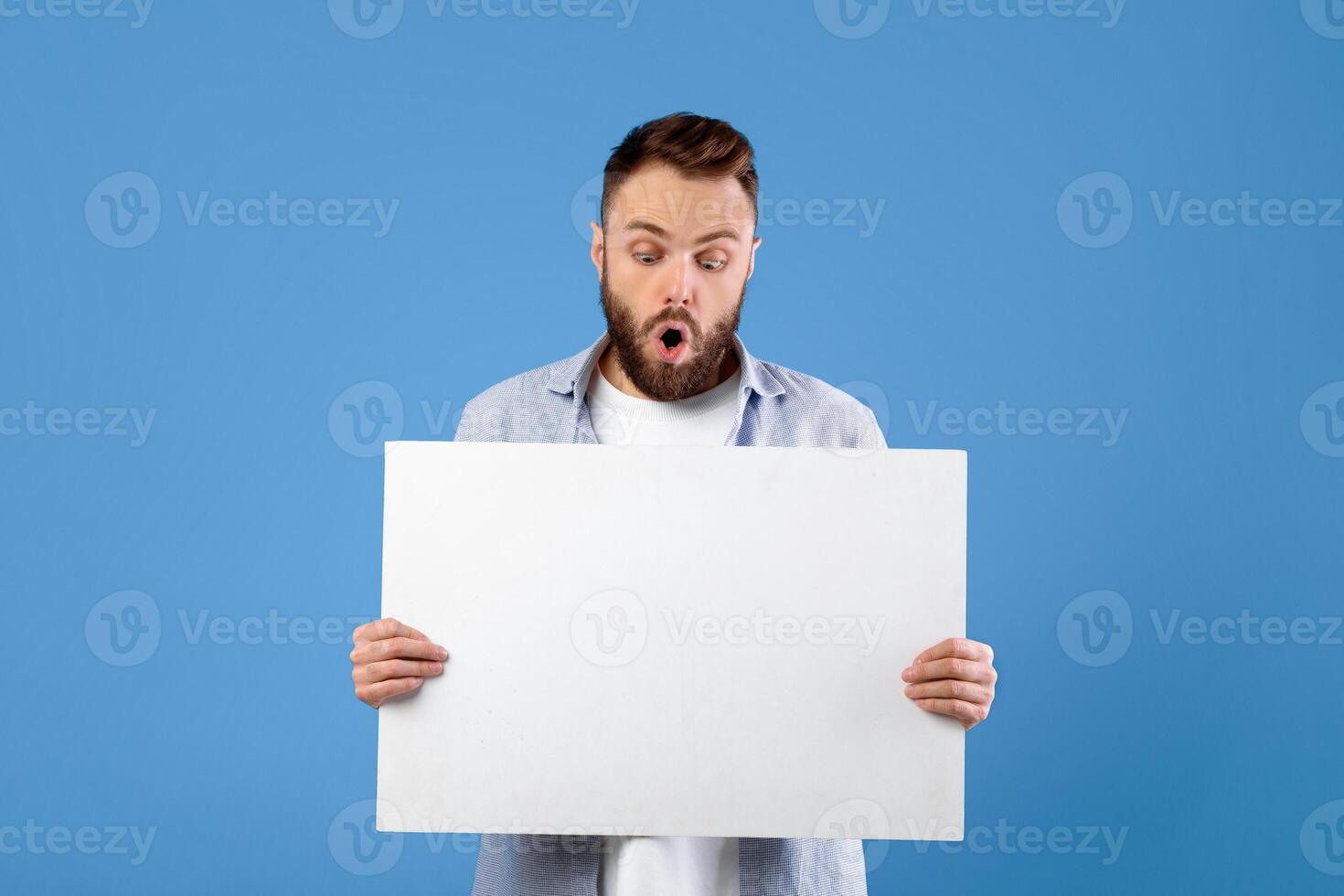 Shocked bearded man holding white empty banner on blue studio background, mockup for your ad or design. Surprised millennial guy with blank poster, hearing unbelievable news, free space photo