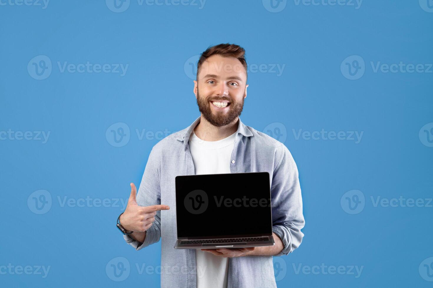 Portrait of happy bearded man holding laptop computer with empty screen over blue studio background, mockup for design. Young guy showing PC with space for your website or advertisement photo