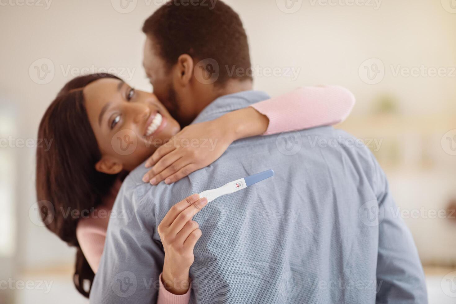 Millennial balck couple excited about positive results of pregnancy test, hugging each other at home. Beautiful expectant young woman embracing her loving husband indoors photo