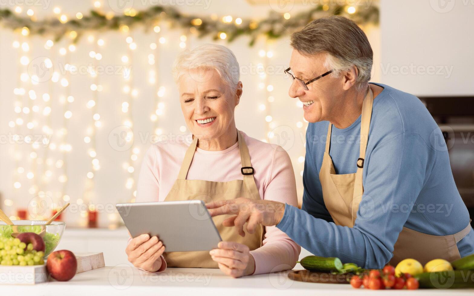 Happy Senior Couple Cooking Using Digital Tablet Searching Christmas Recipes Preparing Dinner In Kitchen At Home. Cooking Application, Family Holidays Concept. Empty Space photo