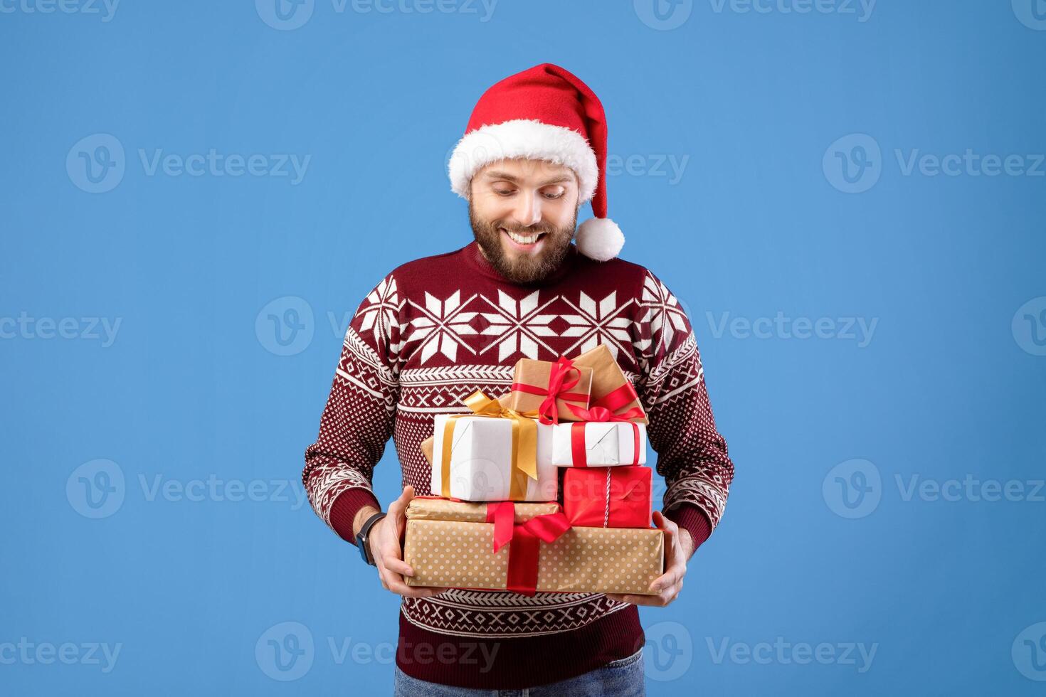 Portrait of happy Caucasian guy in Xmas sweater and Santa hat holding pile of holiday gifts on blue studio background. Handsome young man with stack of presents celebrating Christmas or New Year photo