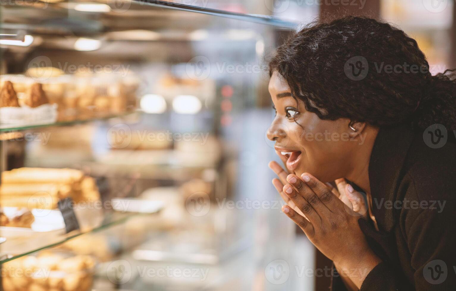 Awesome cakes and sweet tooth concept. Surprised amazed happy african american millennial lady with open mouth looks at shop window with desserts in modern candy shop, close up, copy space photo