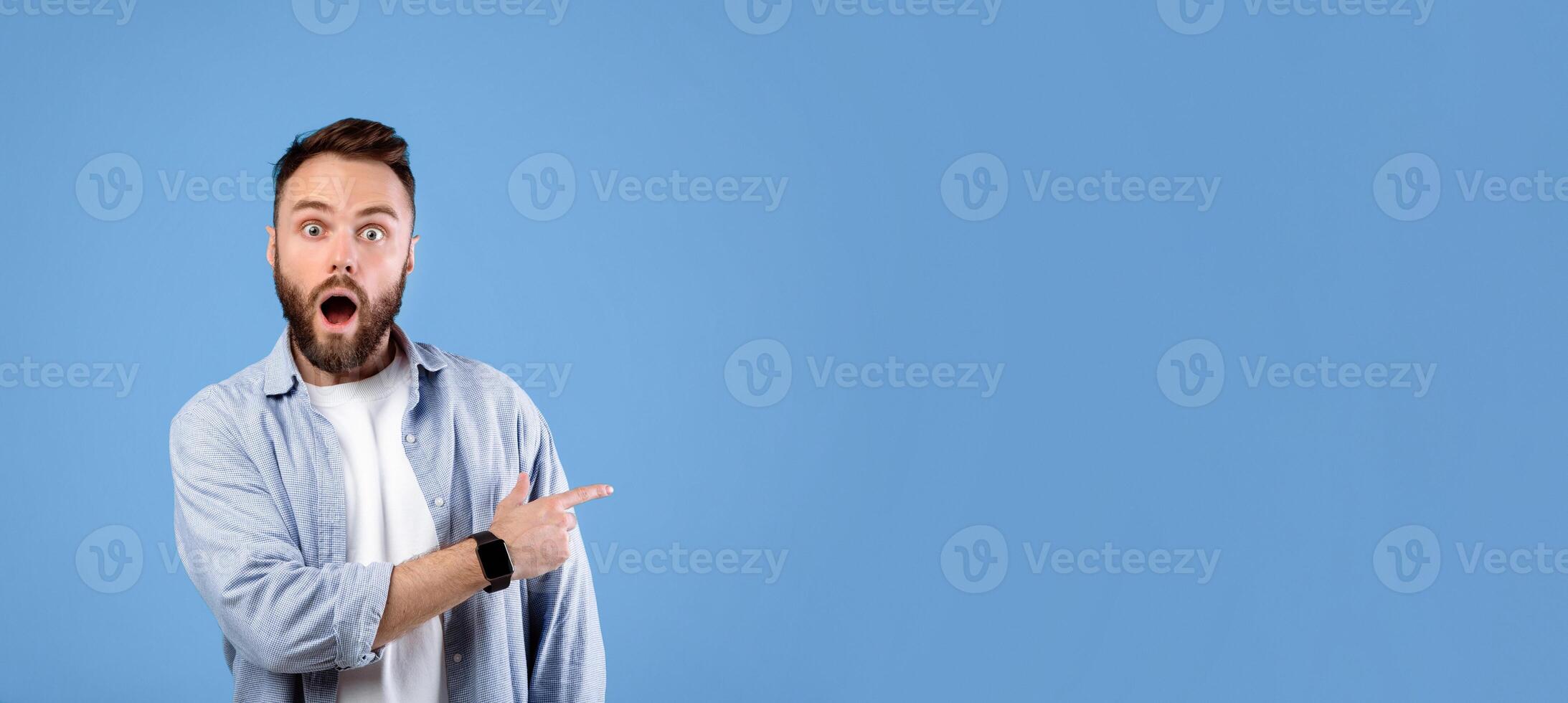 Shocked young guy standing with open mouth, pointing at empty space in disbelief, blue studio background, banner design. Overwhelmed bearded man offering huge sale or promotion photo