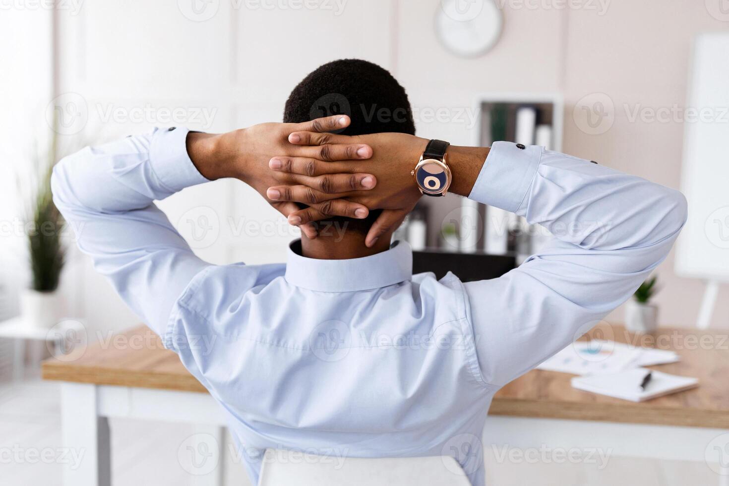 Back view of black businessman in formal shirt sitting at workplace in his office, sitting in chair and leaning back, thinking about something or relaxing during difficult working day, copy space photo