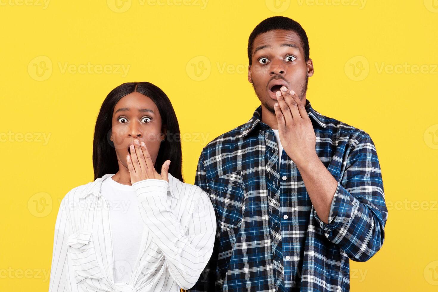 Shocking news, unexpected events. Surprised scared millennial african american female and man with wide open eyes, covering mouth with hand, isolated on yellow background, studio shot, free space photo