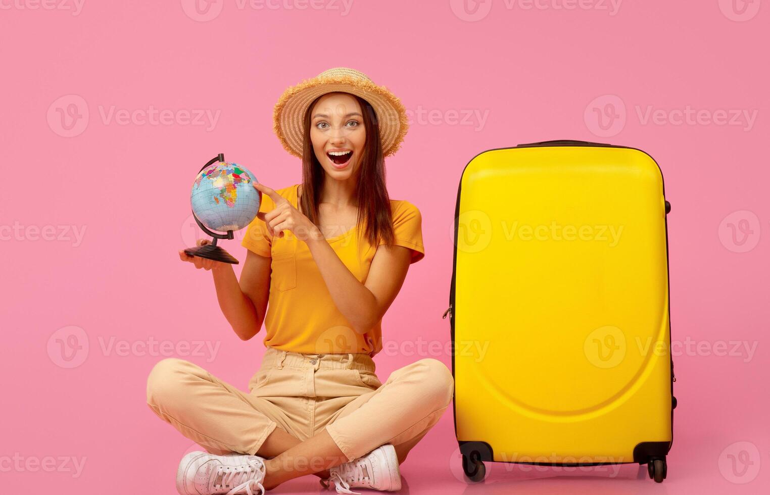 Emotional girl with globe sitting next to yellow luggage over pink background photo