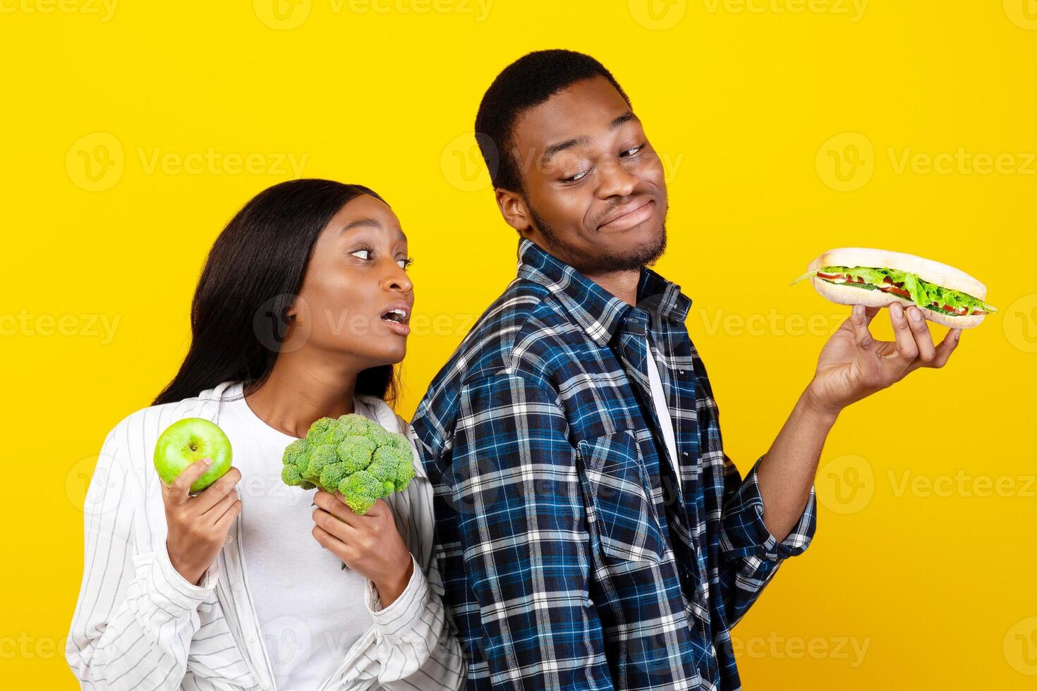 Hunger, envy, vegetarian and healthy eat. Sad young african american wife with broccoli and apple in hands looks at cheerful guy with sandwich, isolated on yellow background, studio shot, free space photo