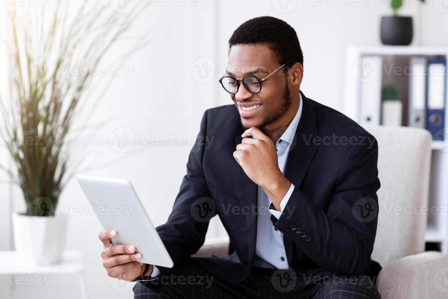 Cheerful black businessman holding digital tablet, office interior, empty space. Smiling african american young entrepreneur using digital pad while working in his office, surfing on internet photo