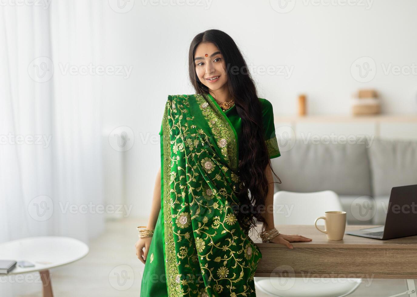 Portrait of beautiful Indian lady in green sari dress leaning on table and smiling at camera, indoors. Attractive young Eastern woman in ethnic clothes and jewelry posing at home photo