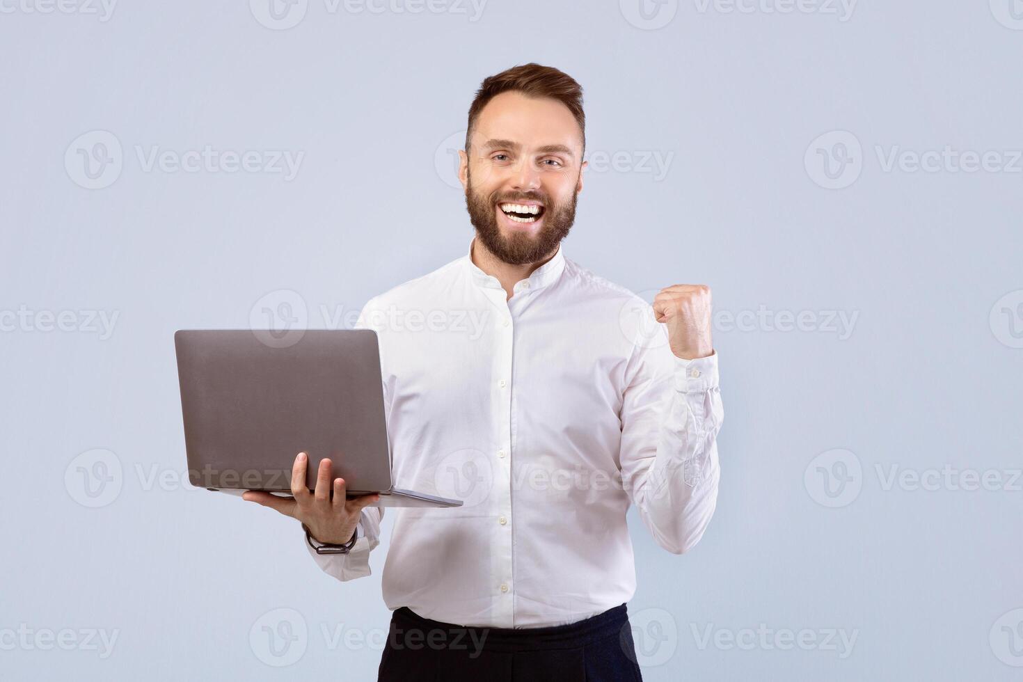 Excited office worker in formal wear holding laptop and making YES gesture over grey studio background. Positive young man working online, celebrating his success or business achievement photo