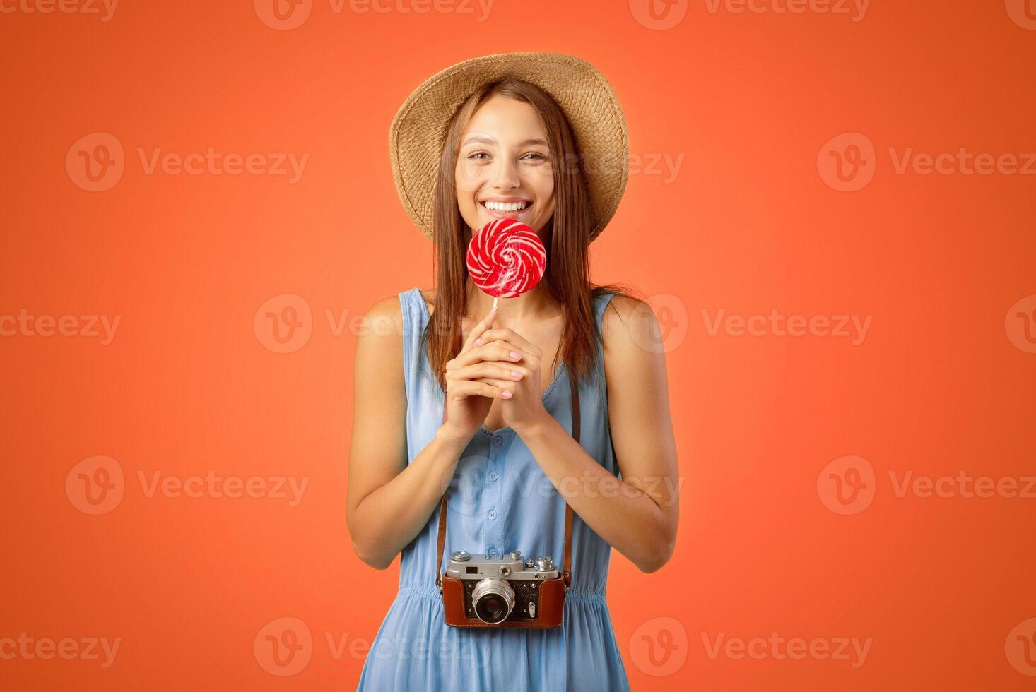 Pretty lady traveller in summer hat with camera eating candy, yellow studio background. Happy young woman with candy smiling at camera, copy space. Joyful female tourist going vacation photo