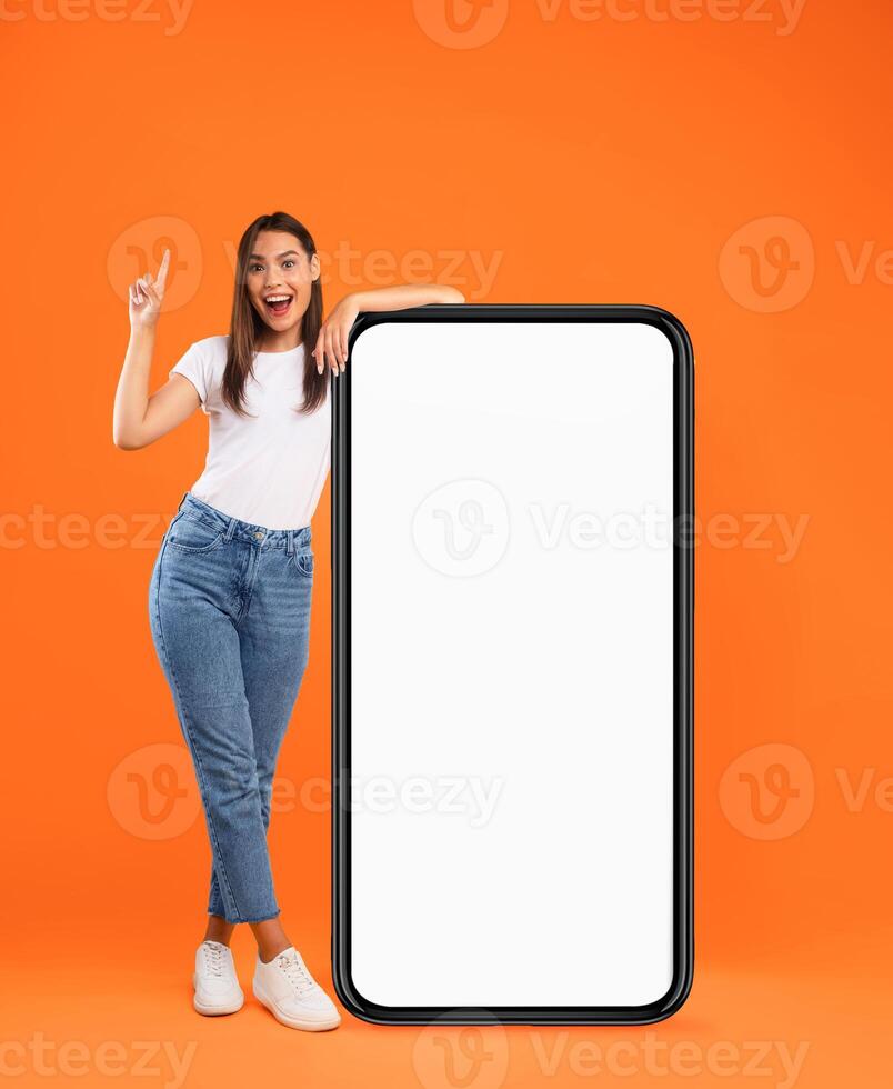 Mobile App Advertisement, Great Offer. Full Body Length Of Excited Lady Pointing Finger Up Leaning On Big Huge White Empty Smartphone Screen Standing On Orange Studio Wall. Cellphone Display Mock Up photo