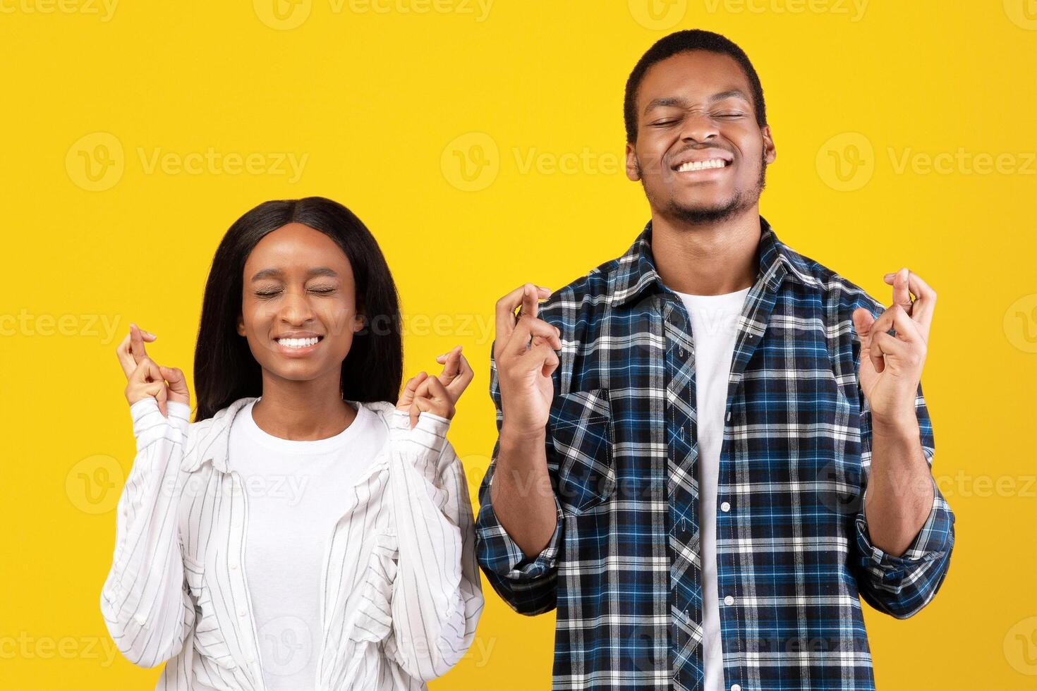 Make a wish, really want a dream come true. Excited millennial african american students man and lady in casual with fingers crossed and closed eyes, isolated on yellow background, empty space photo