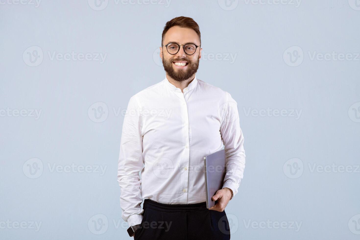 Happy young businessman in formal clothes and glasses holding laptop computer on grey studio background. Positive millennial entrepreneur with pc looking at camera and smiling photo