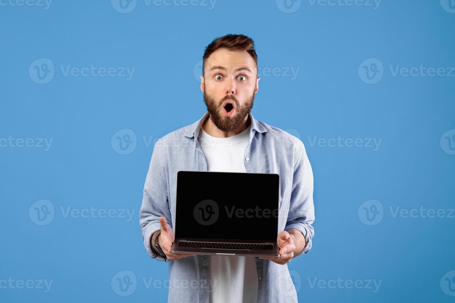Shocked bearded guy showing laptop with blank screen on blue studio background, mockup for your website. Millennial man demonstrating PC with space for your online advertisement photo