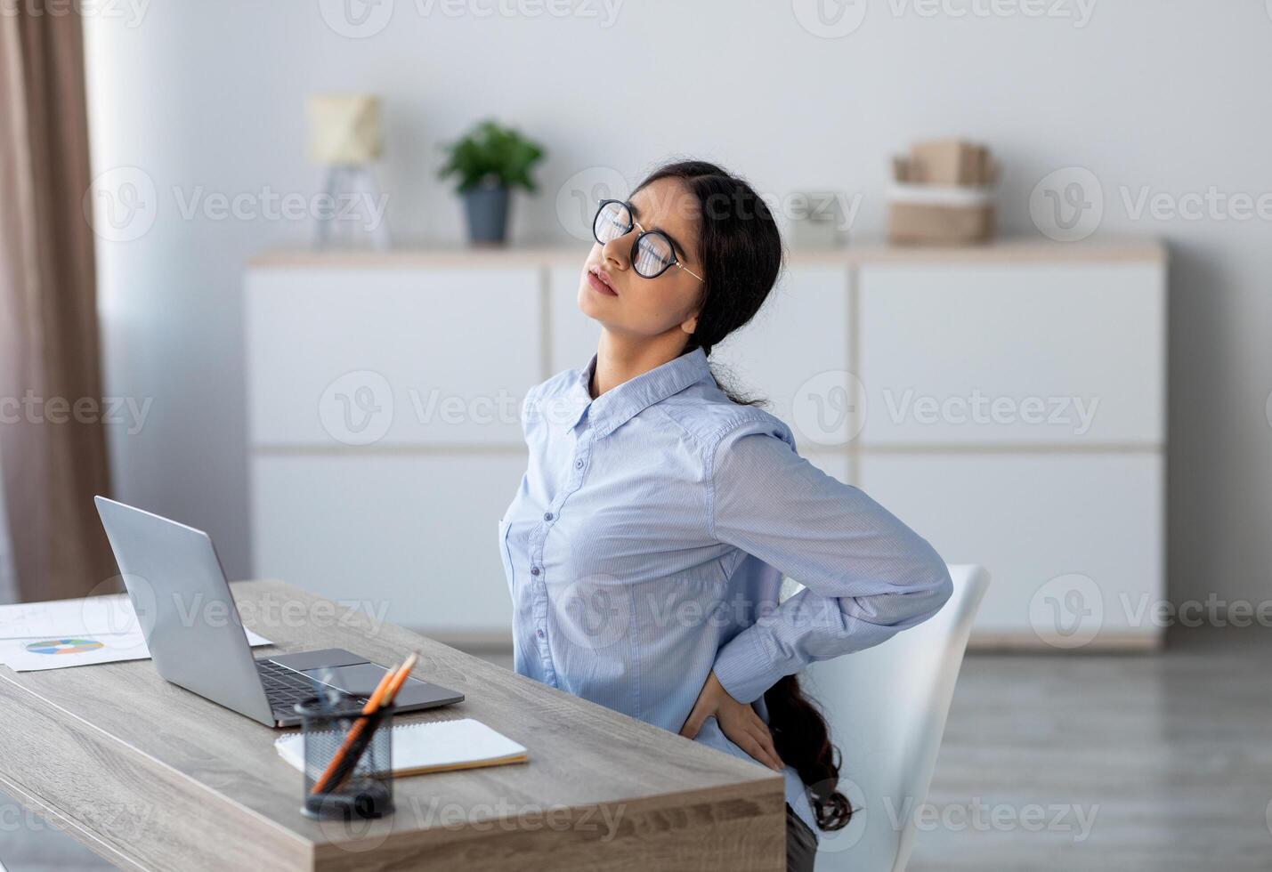 Tired upset young indian business woman suffering from back pain, muscle spasm at workplace with laptop in office or home interior. Fatigue, deadline, pain and incorrect posture, work at computer photo