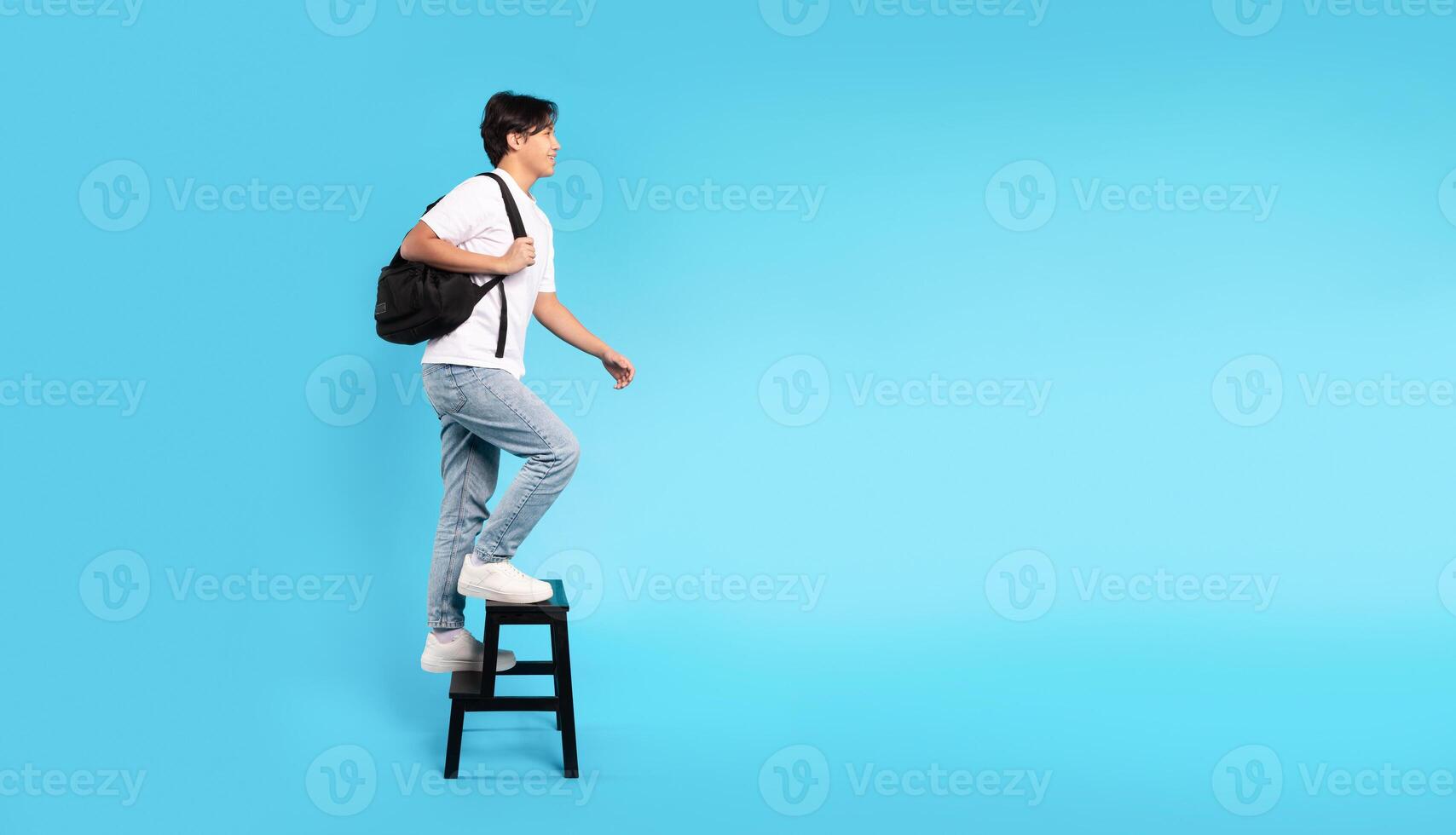 Education And Self Growth. Happy Korean Teen Guy Walking Up Stairs On Ladder Looking Aside Posing Holding Backpack Over Blue Studio Background. Panorama With Copy Space, Side View Shot photo
