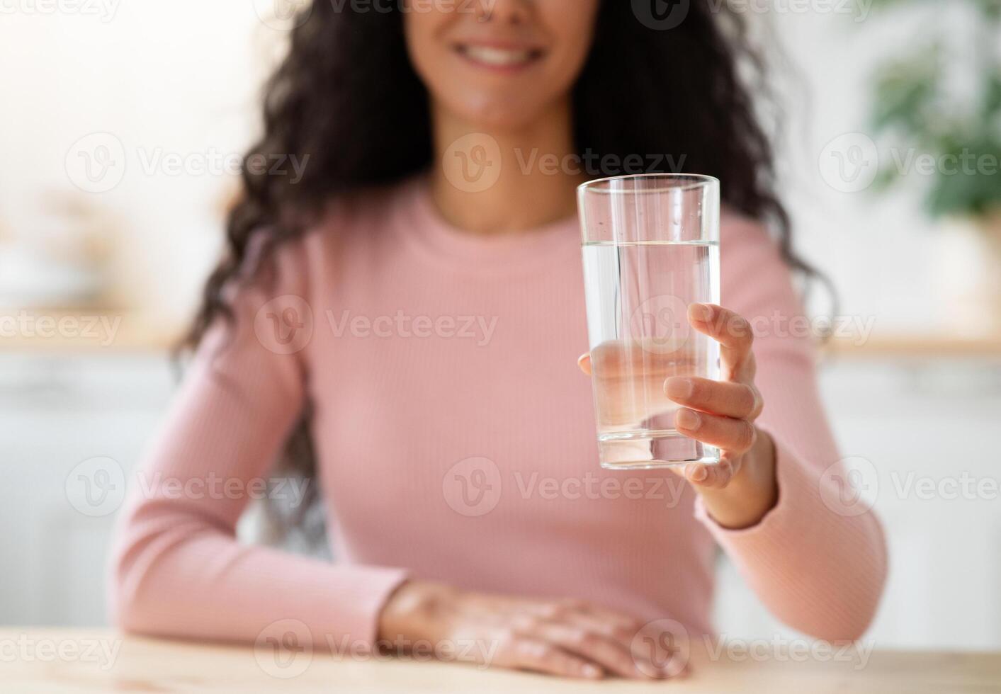 Healthy Drink. Unrecognizable Smiling Lady Offering Glass With Mineral Water At Camera While Sitting At Table In Kitchen, Brunette Woman Drinking Aqua At Home, Closeup Shot With Selective Focus photo