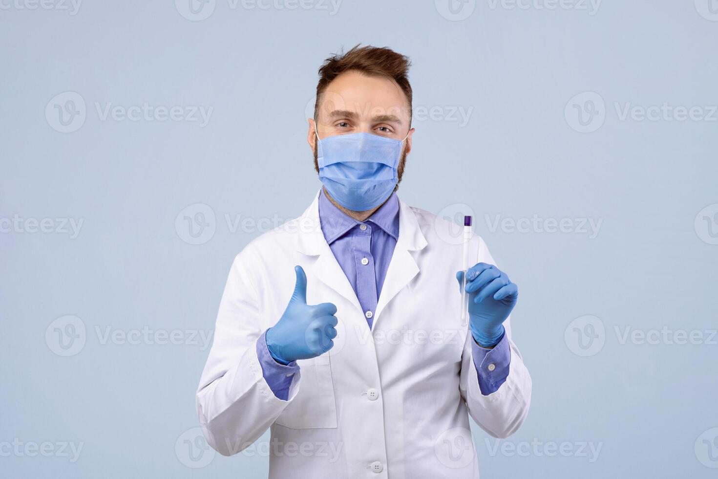 Portrait of young doctor in mask and gloves holding covid-19 PCR test tube and showing thumb up gesture on grey studio background. Medical worker recommending coronavirus diagnostic procedure photo