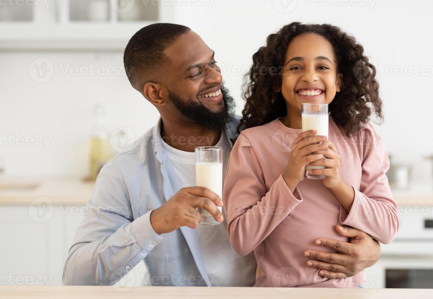 Nutrition Concept. Portrait of smiling black girl holding glass and drinking milk with her father in the kitchen. Happy dad hugging his cute curly daughter, having healthy meal together photo