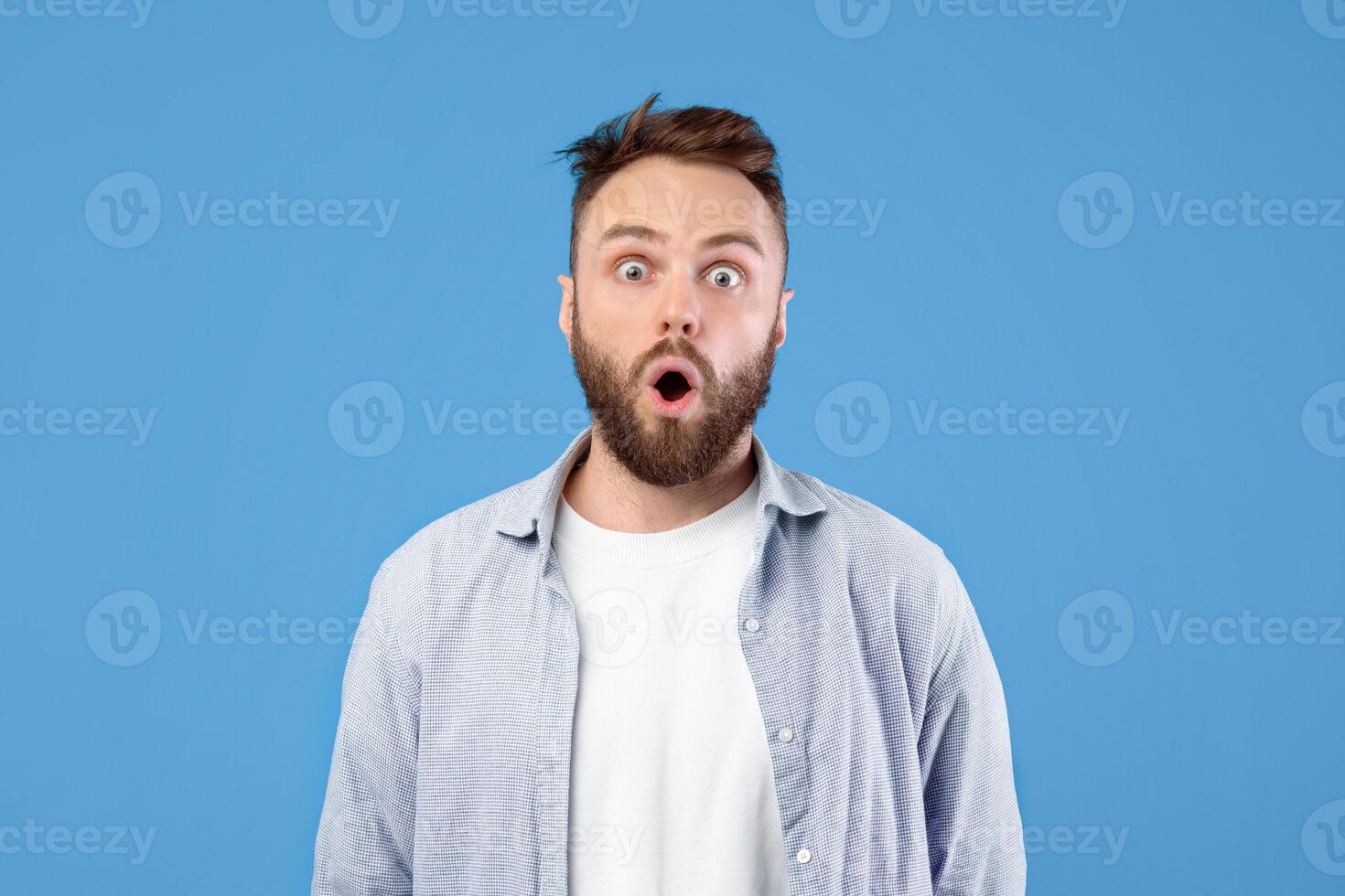Portrait of stunned young guy opening mouth in shock on blue studio background. Amazed millennial man in casual clothes having shocked face expression, cannot believe his luck, learning amazing news photo